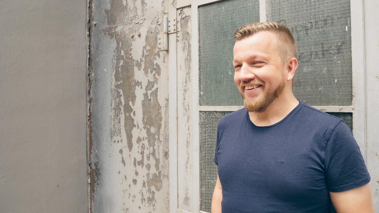 Jakob Klimkait steht lachend vor einem offensichtlich älteren Gebäude. Die Farbe blättert von Wänden und Türen. Er trägt ein T-Shirt.